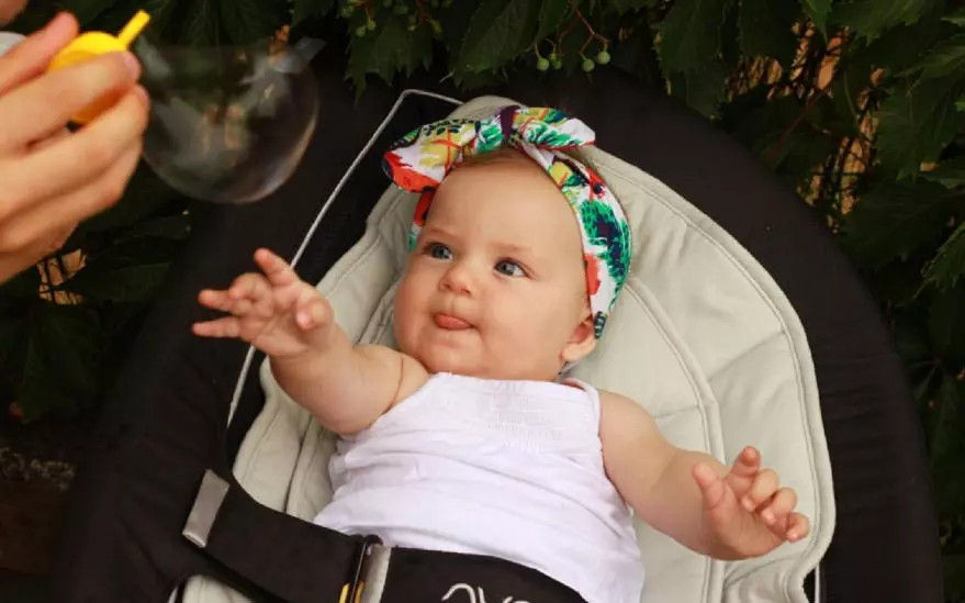 Un bebe portant un bandeau colore et une tenue blanche est assis dans une poussette, tendant la main vers une bulle soufflee a proximite. Le decor presente un feuillage vert luxuriant en arriere-plan, creant une atmosphere naturelle et ludique. Le bebe semble interesse et curieux, interagissant avec la bulle.