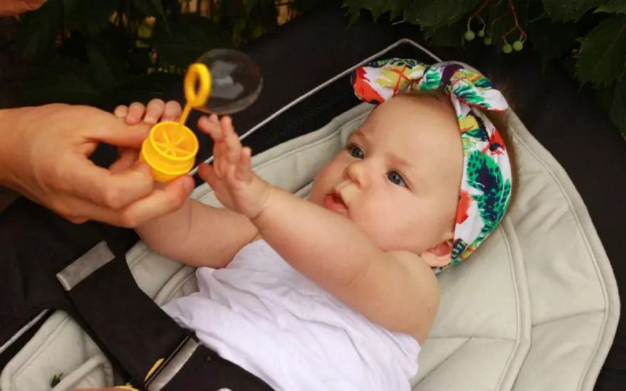 Un bebe portant un bandeau floral colore est assis dans une poussette et joue avec un jouet a bulles jaune. Le decor semble etre en exterieur, entoure de verdure. La scene degage une atmosphere ludique et joyeuse gr�ce a des couleurs vives et des elements naturels.