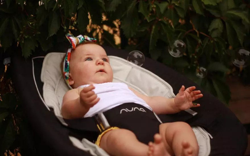 Un bebe est assis dans un siege bebe de marque Nuna a l'exterieur, entoure de verdure. L'enfant porte une tenue blanche et un bandeau colore. Des bulles transparentes flottent autour de lui, ajoutant une touche ludique a la scene. Le decor suggere un environnement detendu et naturel.