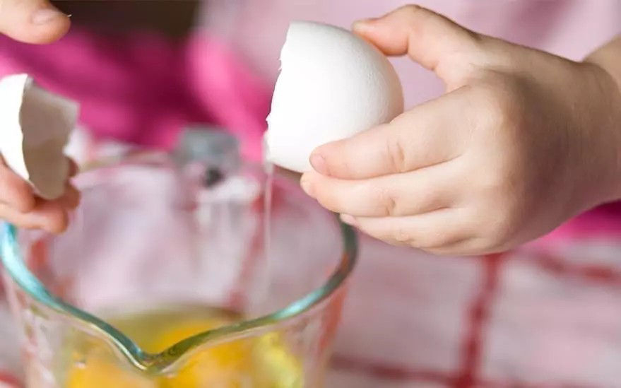 On voit un jeune enfant casser un �uf dans un bol en verre transparent. Le decor semble etre une cuisine avec une surface a carreaux rouges et blancs en arriere-plan. L'accent est mis sur les mains de l'enfant et l'�uf, dont le jaune est visible dans le bol. L'image degage une impression d'apprentissage ou d'activite culinaire ludique.