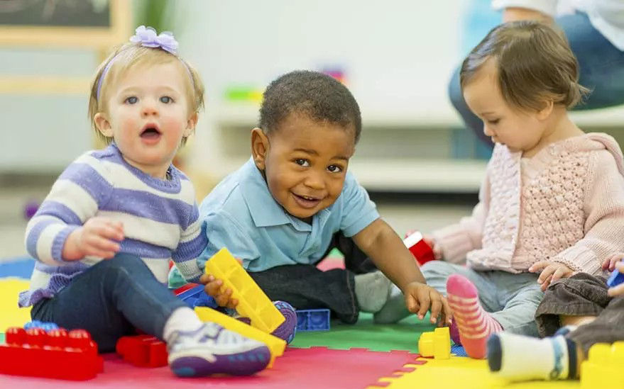 Un groupe de jeunes enfants assis sur un tapis en mousse aux couleurs vives, jouant avec de gros blocs de construction colores. Le decor semble etre une salle de jeux interieure ou une garderie d' . Les enfants sont vetus de tenues decontractees et la scene degage une atmosphere ludique et joyeuse.