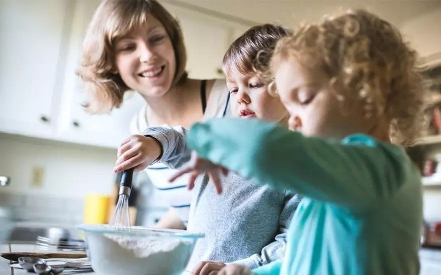 Une famille est en train de cuisiner dans une cuisine lumineuse. Un saladier rempli de farine est en train d'etre battu, avec des ustensiles et des ingredients visibles en arriere-plan. La scene degage une atmosphere chaleureuse et engageante de convivialite et de cuisine maison.