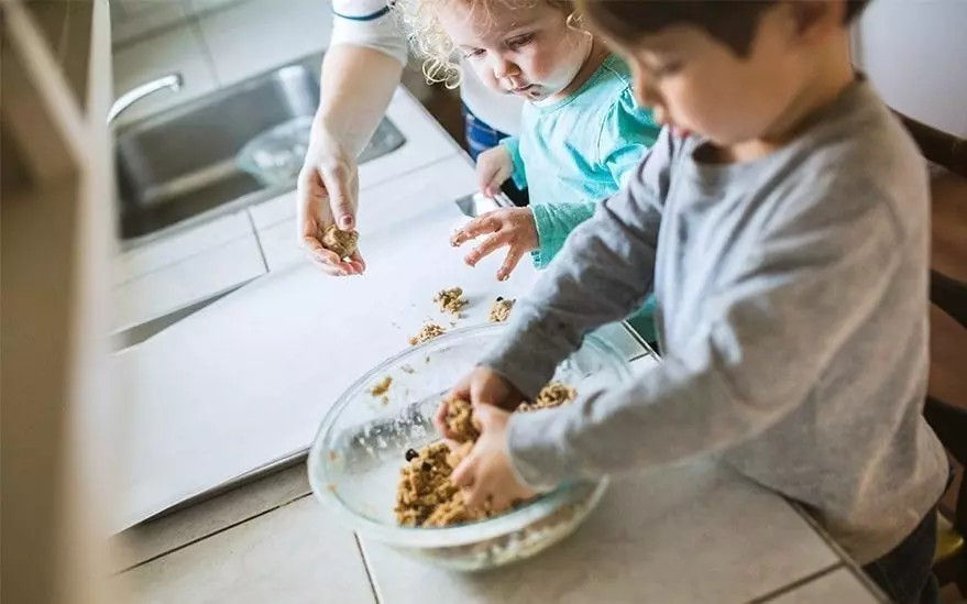 On voit deux enfants preparer de la p�te a biscuits dans un saladier en verre sur le plan de travail de la cuisine. L'un des enfants fa�onne la p�te tandis que l'autre lui tend la main pour l'aider. La scene se deroule dans une cuisine lumineuse avec un evier visible a l'arriere-plan. L'activite degage une atmosphere ludique et collaborative.