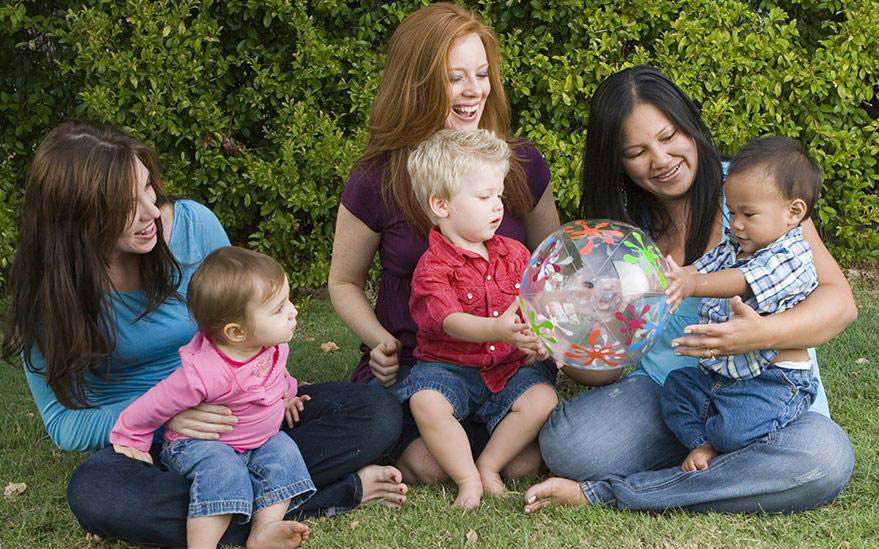 Un groupe de femmes et de jeunes enfants sont assis sur l'herbe a l'exterieur. Les enfants jouent avec un ballon gonflable colore a motifs floraux. La scene est entouree d'une vegetation luxuriante, creant une atmosphere decontractee et joyeuse. L'activite met en avant les liens familiaux et les jeux en plein air.