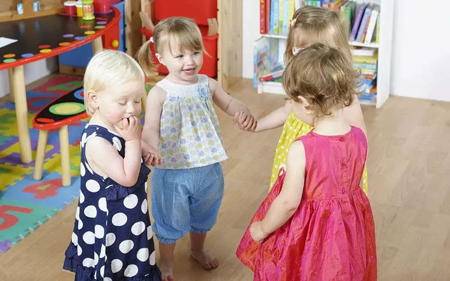 On voit un groupe de quatre enfants en bas �ge se tenant par la main et interagissant dans une salle de jeux interieure bien eclairee. Le decor comprend des meubles colores, un tapis de jeu et une bibliotheque remplie de livres. Les enfants sont vetus de tenues aux couleurs vives, notamment a pois et a motifs floraux, creant une atmosphere joyeuse et animee.