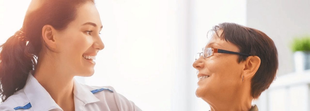 Eine junge Frau in weißer Kleidung lächelt eine ältere Frau mit Brille in einem hellen Raum freundlich an.