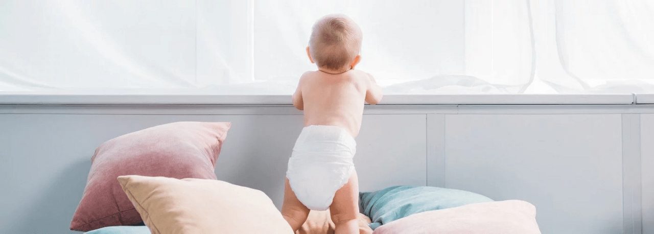 Ein Baby in Windel steht zwischen bunten Kissen an einem Fenster und blickt nach draußen in einen hellen Raum.