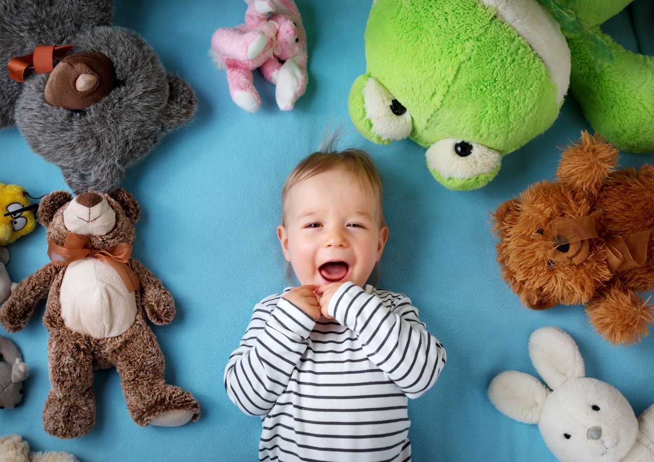 Happy one year old boy lying with many plush toys on blue blanket