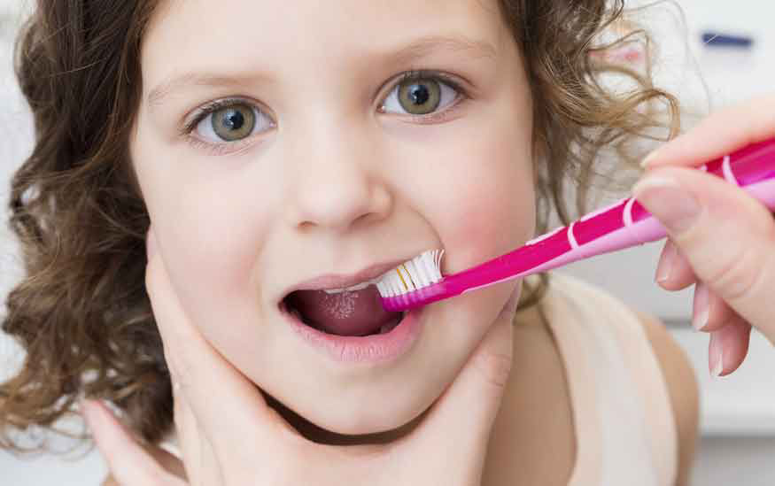 Gros plan sur une personne recevant des soins dentaires a l'aide d'une brosse a dents rose. Le decor semble etre interieur, peut-etre une salle de bain ou un cabinet dentaire. La brosse a dents est d'un rose vif avec des accents blancs, ajoutant un element visuel ludique. L'accent est mis sur l'hygiene bucco-dentaire et les soins personnels.