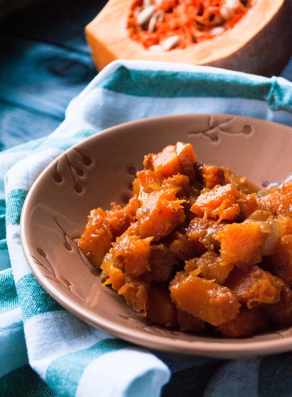 A bowl of cooked pumpkin cubes served in a beige ceramic dish with a floral pattern. The setting features a rustic blue wooden surface and a teal-striped cloth napkin. A halved pumpkin with visible seeds is placed in the background, adding to the autumnal theme.