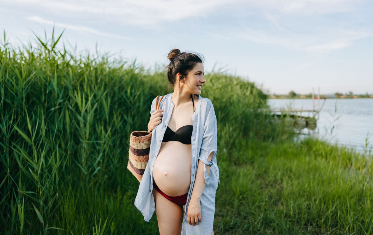 Une femme enceinte se tient dans un cadre exterieur herbeux pres d un plan d eau. Elle porte un haut de bikini noir, un bas de bikini rouge et une chemise ouverte bleu clair. Elle porte un sac en paille tressee et est entouree de grands roseaux verts sous un ciel clair.