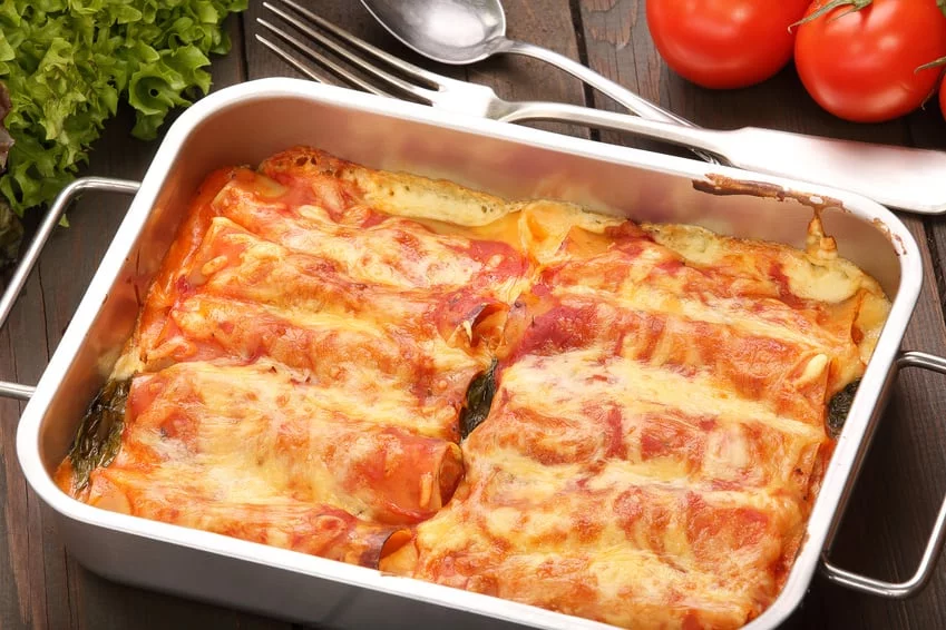 A freshly baked lasagna with melted cheese and tomato sauce served in a white rectangular pan. The dish is placed on a wooden table alongside fresh tomatoes, lettuce, and cutlery. The vibrant colors of the ingredients enhance the appetizing presentation.
