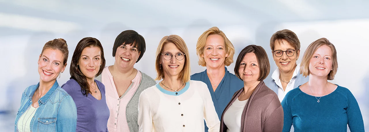 Acht Frauen unterschiedlichen Alters stehen nebeneinander und lächeln vor einem unscharfen, hellen Hintergrund.