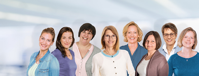 Acht Frauen stehen nebeneinander und lächeln in die Kamera, im Hintergrund ist unscharf ein modernes Büro zu erkennen.