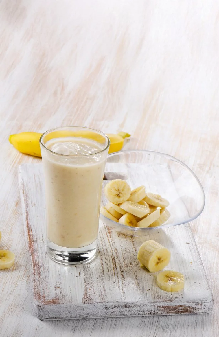 A glass of creamy banana smoothie placed on a rustic wooden surface. Fresh banana slices are arranged in a clear bowl beside a whole banana. The setting features a light, natural palette with a casual and wholesome vibe.