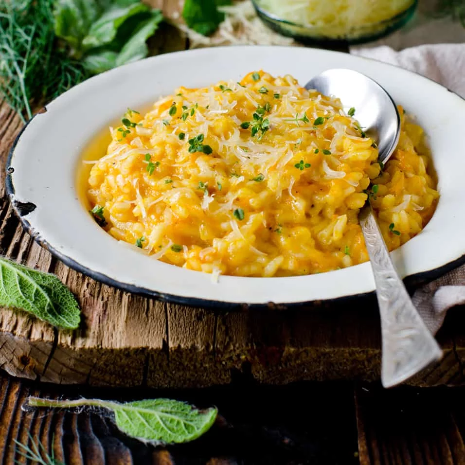 A bowl of creamy pumpkin risotto garnished with fresh herbs and shredded cheese sits on a rustic wooden surface. The dish is served in a white enamel bowl with a silver spoon, surrounded by fresh sage leaves and greenery. Warm tones and natural textures create a cozy, autumn-inspired mood.