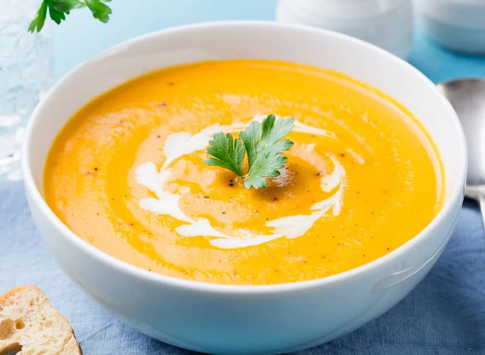 A vibrant bowl of creamy pumpkin soup garnished with fresh parsley and a swirl of cream. The dish is set on a light blue tablecloth with a slice of bread nearby, creating a cozy dining atmosphere. The warm orange tones of the soup contrast beautifully with the cool blue background.