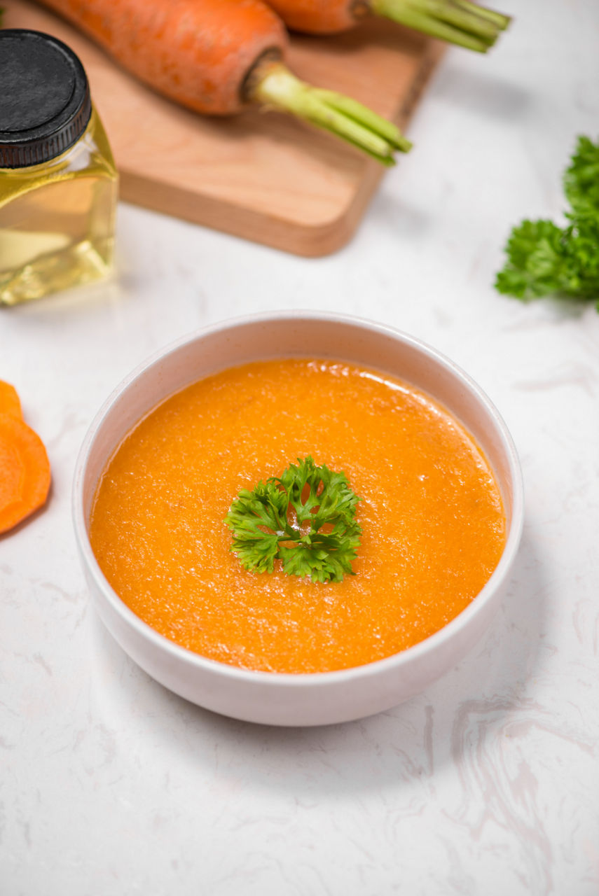 A bowl of vibrant orange carrot soup is garnished with fresh parsley, creating a visually appealing dish. The setting includes raw carrots, a bottle of oil, and parsley on a white marble surface, emphasizing freshness and natural ingredients. The soup's smooth texture and bright color evoke a warm and healthy meal.