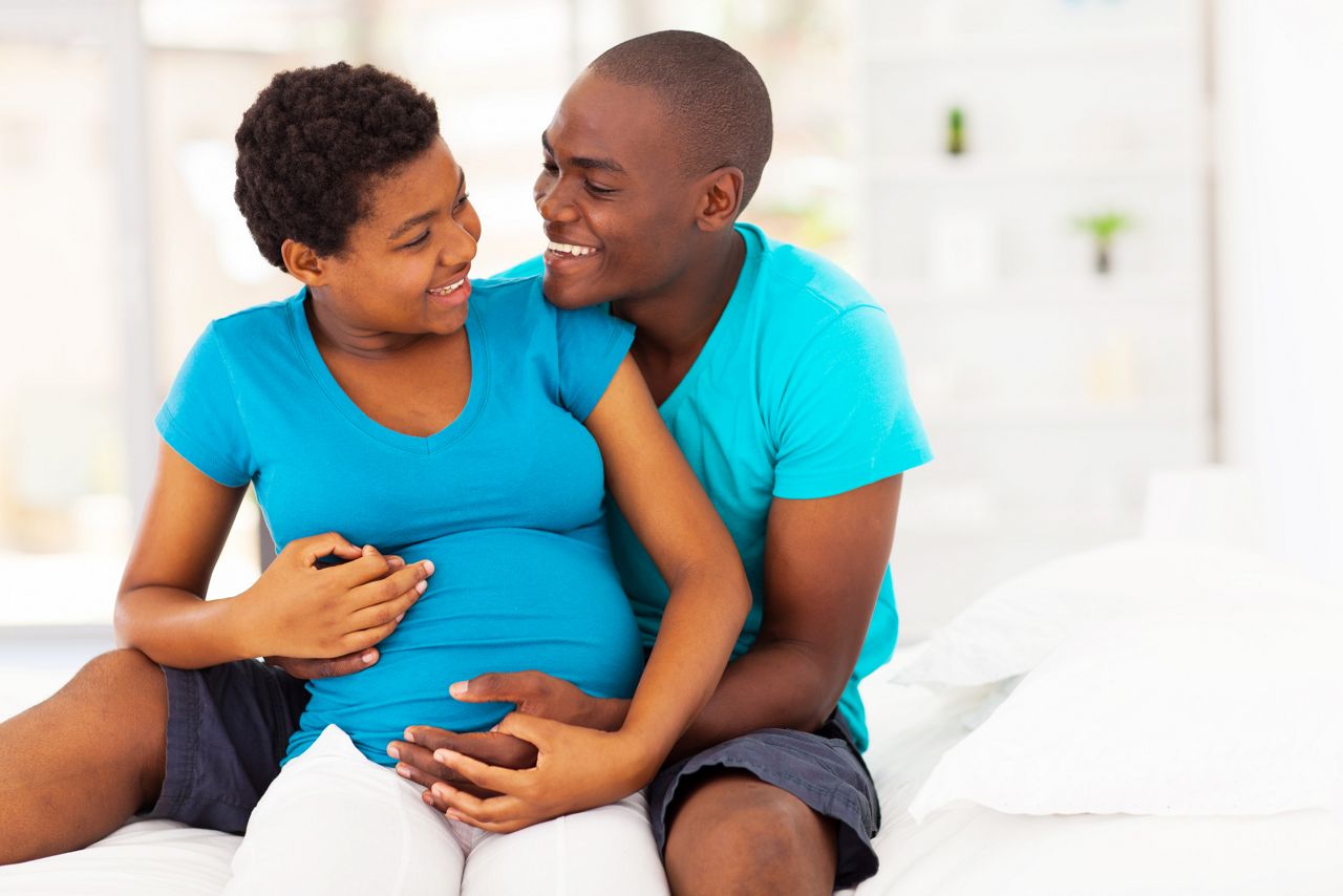 happy african american pregnant woman and husband hugging on bed at home