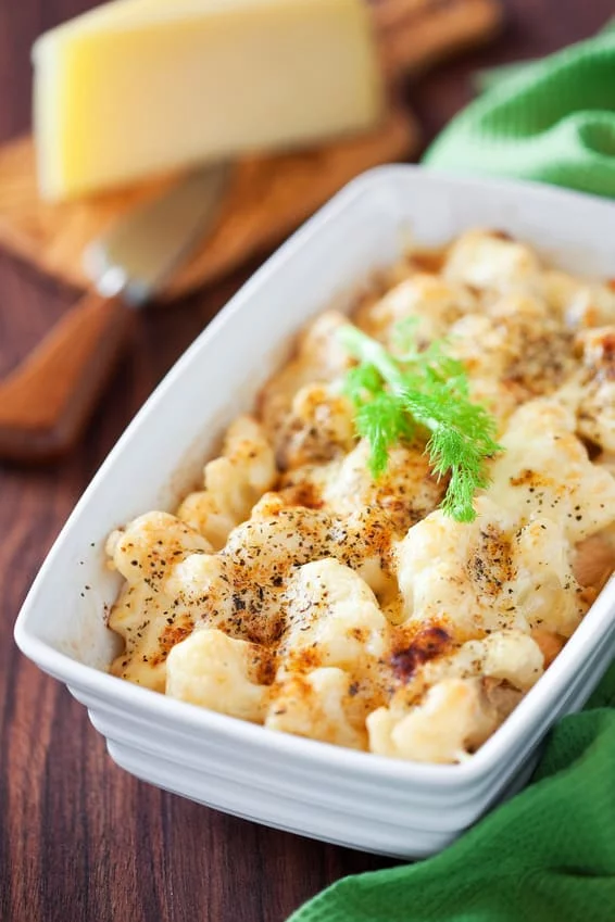 A close-up of baked cheesy pasta served in a white rectangular dish, garnished with fresh herbs. The dish is placed on a wooden table alongside a block of cheese and a green cloth napkin. The golden-brown crust and creamy texture highlight the comfort food appeal.