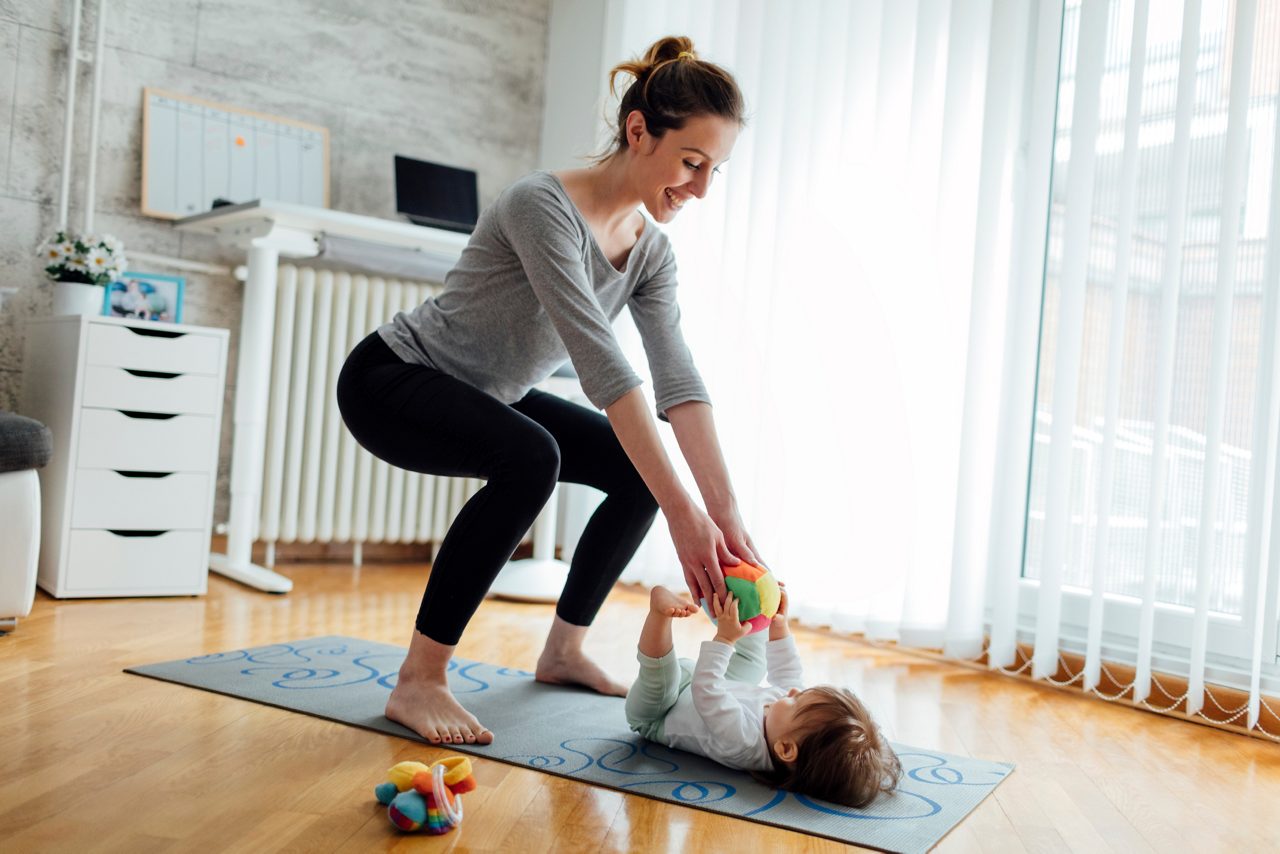 Shot of an attractive young woman with her baby girl while doing exercise. Woman working squats and baby lying on exercise mat. They exercise in a living room. Mother playing with her baby while exercise.