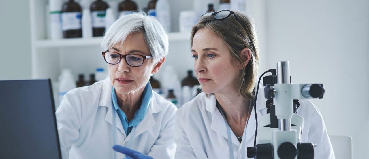 Zwei Wissenschaftlerinnen in Laborkitteln analysieren Daten am Laptop neben einem Mikroskop in einem Laborraum.