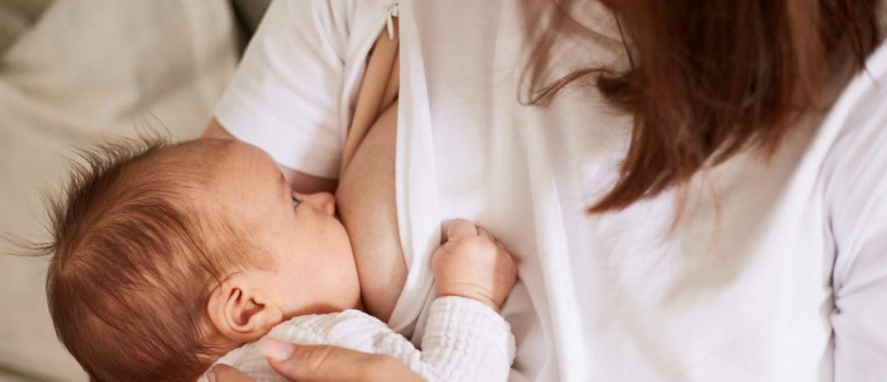 Ein Baby wird im Arm gehalten und gestillt, die Mutter trägt ein weißes Oberteil, im Hintergrund ist ein Sofa erkennbar.