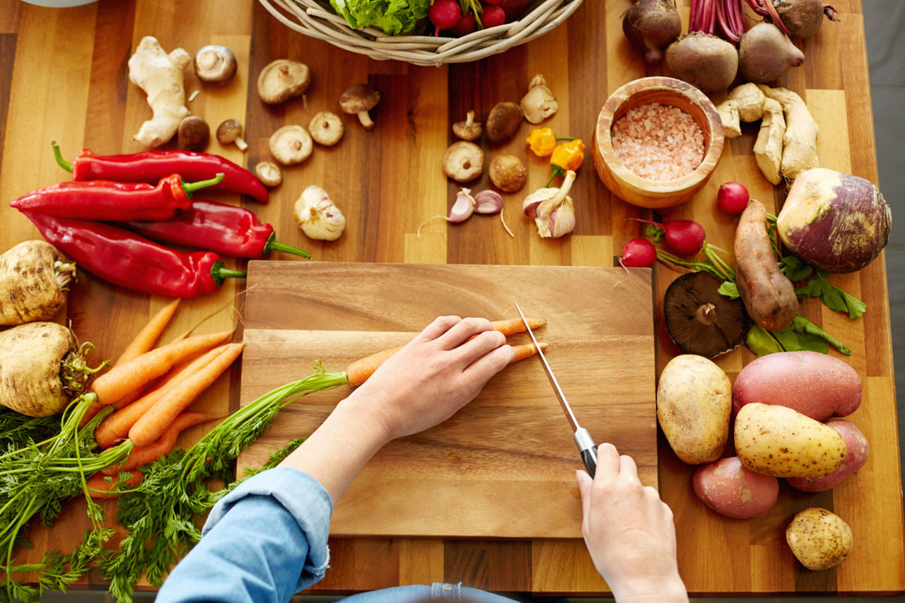 Point of view shot of female hands cutting carrots on chopping board. Various fresh vegetables are on wooden table. Woman is cooking food. She is domestic kitchen.