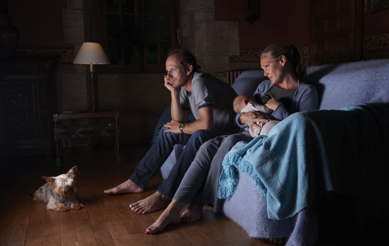 Une scene d interieur chaleureuse mettant en scene un couple assis sur un canape avec un bebe, eclaire par un eclairage chaud. Un petit chien est assis attentivement sur le plancher en bois a proximite. Le cadre comprend une douce couverture bleue, une lampe et un decor rustique, creant une atmosphere calme et intime.