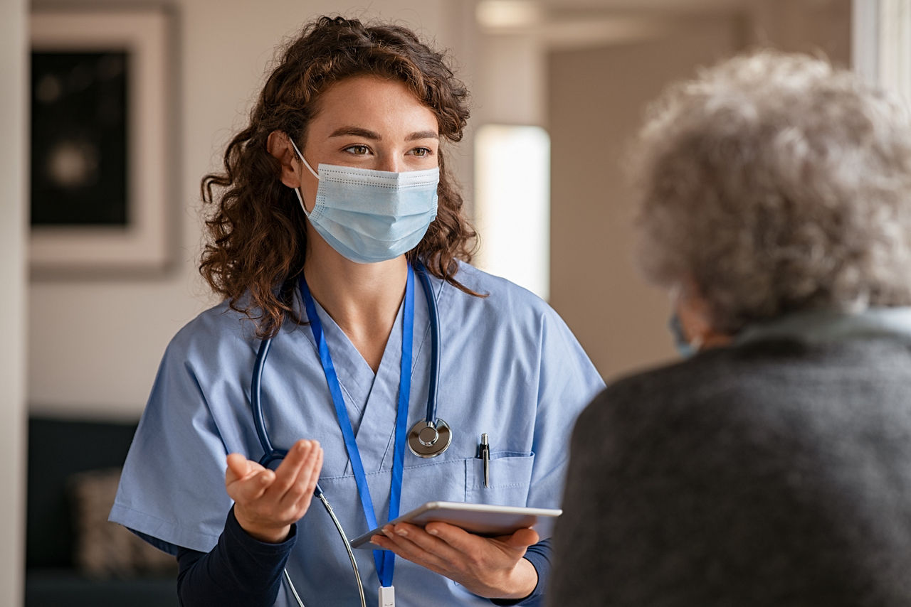 Female doctor holding digital tablet and wearing safety protective mask while talking to senior woman about medical report during a home visit. General pratictioner and senior woman wearing facemasks during coronavirus and flu outbreak. Caring nurse supporting and cheering up senior patient during home visit during covid-19 pandemic.