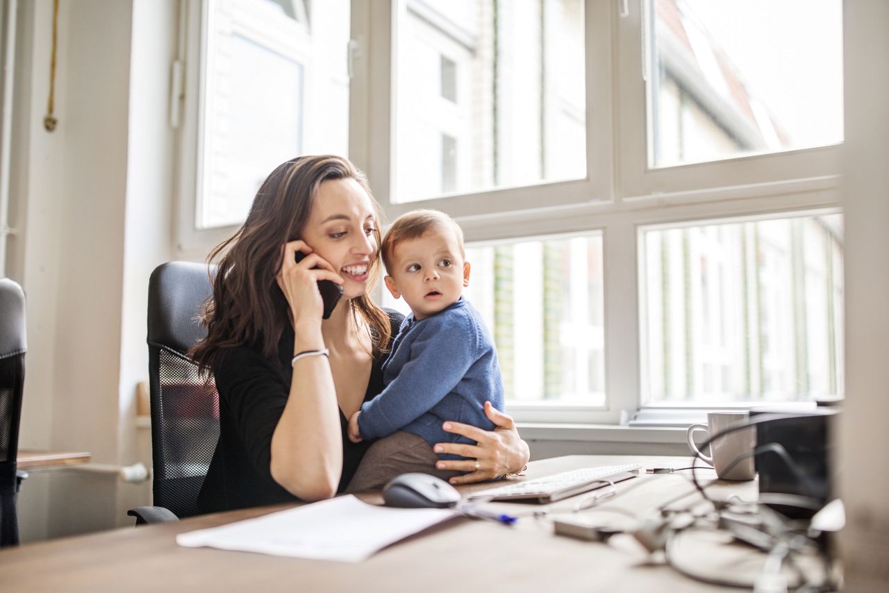 Young woman sitting at table with her son talking on mobile phone in office. Single mother with son working in office.