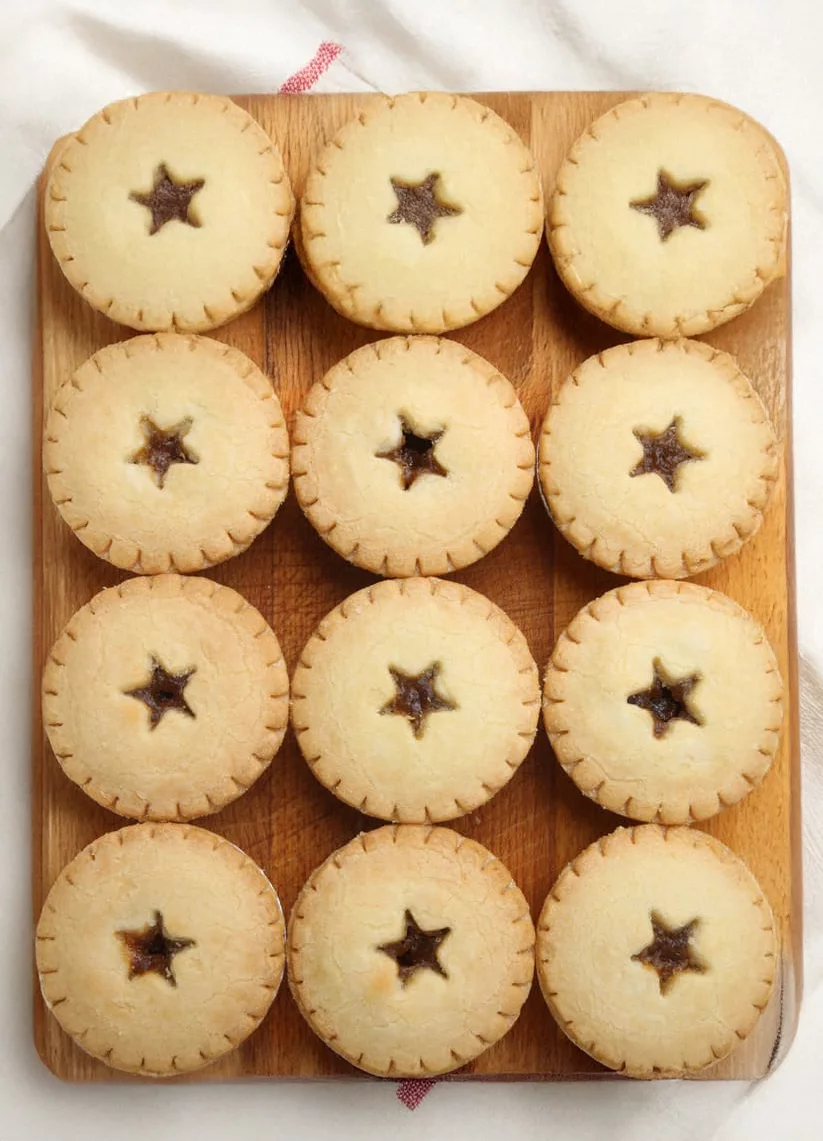 A neatly arranged set of twelve pastries with star-shaped cutouts, placed on a wooden tray. The pastries appear to be filled with a dark filling, possibly fruit-based. The setting includes a soft white fabric background with a hint of red stitching visible. The overall mood is warm and inviting, ideal for dessert or snack imagery.