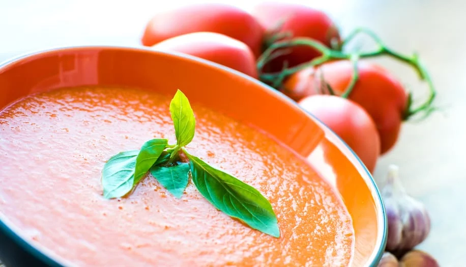A vibrant bowl of tomato soup served in an orange dish, topped with fresh basil leaves. The setting includes ripe tomatoes and garlic in the background, emphasizing a fresh and natural theme. The image highlights the rich red color of the soup and the contrasting green garnish.