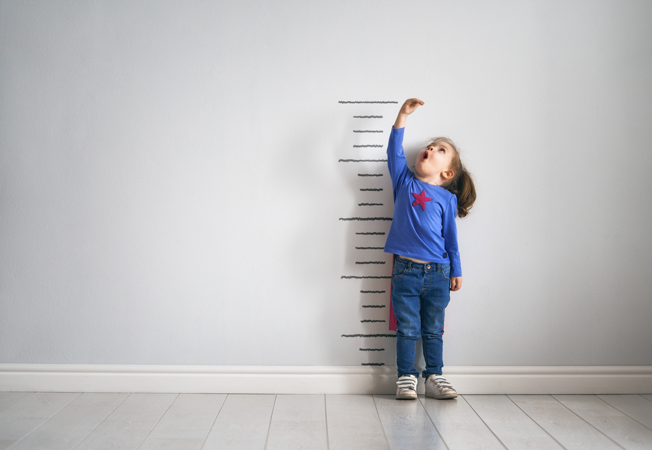 Little child is playing superhero. Kid is measuring the growth on the background of wall. Girl power concept.