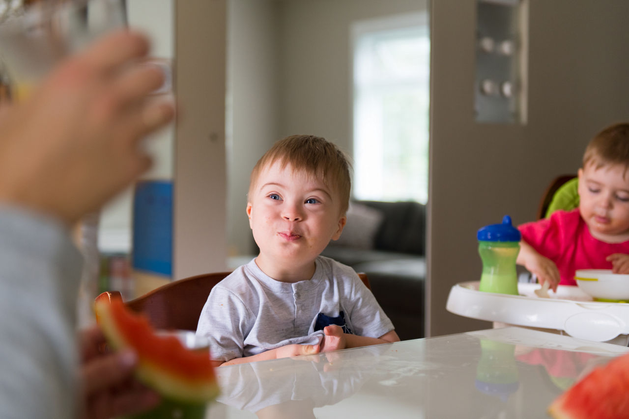 Down's syndrome sweet boy eating breakfast. He has blue eyes and blond hair. He is sitting at the table in the dining room with his 2 years old brother.  They wear pyjama short and t-shirts. The pretty boy is looking his mother. The color and horizontal Photo was taken in Quebec Canada.