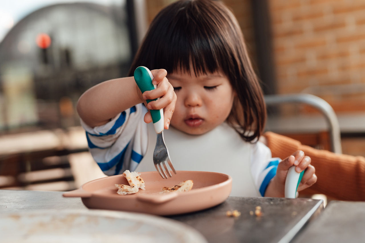 Little Asian girl eating cheesy pizza with a fork at the restaurant. Toddler learning to use utensils. Eating out with kids.