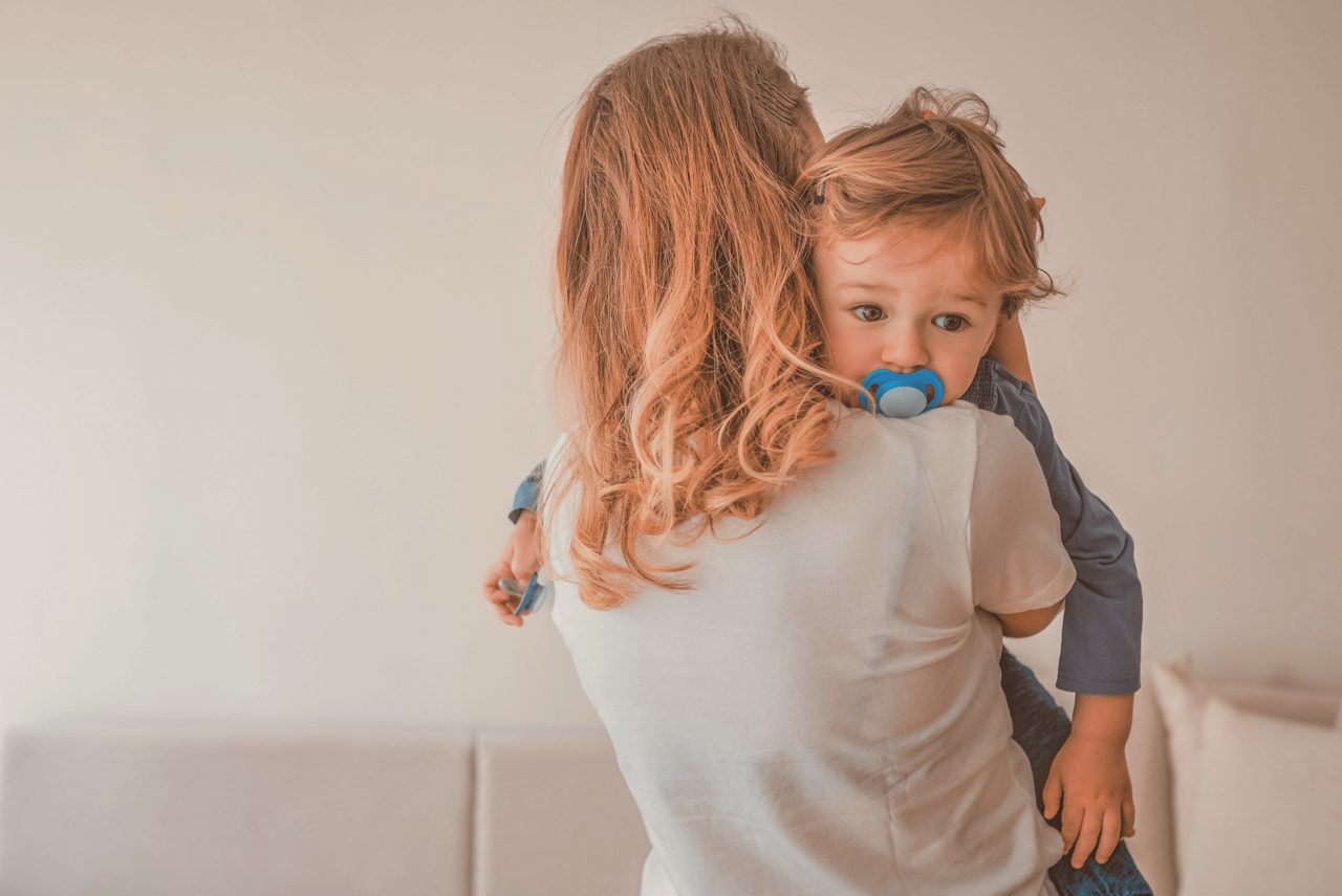 Eine Frau hält ein kleines Kind mit Schnuller auf dem Arm, im Hintergrund steht ein helles Sofa in einem Wohnzimmer.