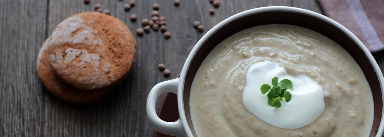 Eine Schale cremige Suppe mit einem Klecks Creme und Kräutern steht auf Holztisch, daneben liegen zwei braune Kekse.