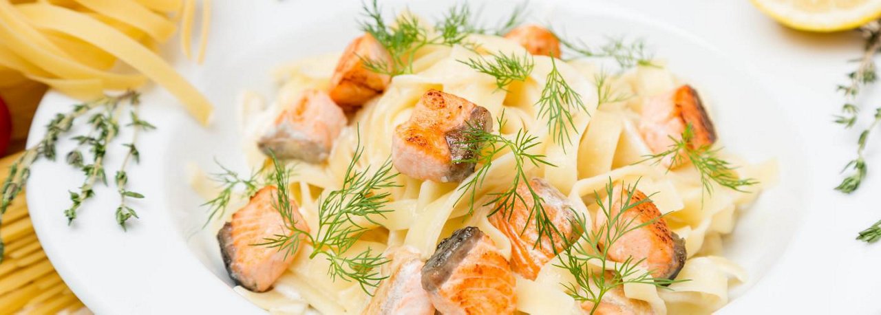 Tagliatelle mit Lachsfilet und frischem Dill auf weißem Teller, daneben rohe Nudeln und Kräuter auf hellem Hintergrund.