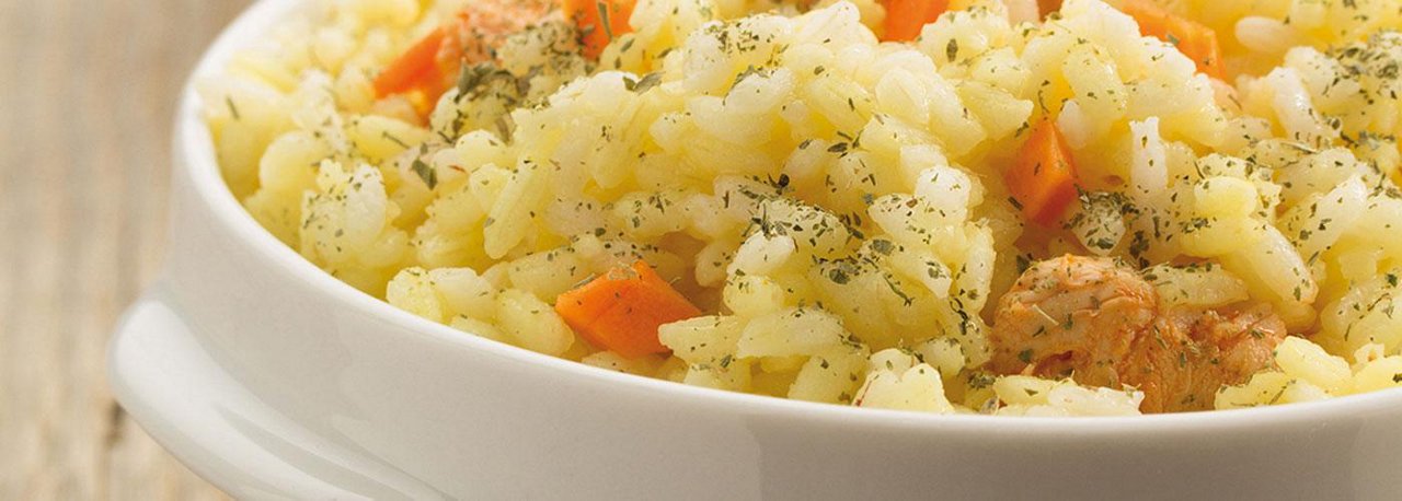 Ein weißer Teller ist gefüllt mit Reis, kleinen Karottenstücken, Fleischwürfeln und Kräutern auf einem Holztisch.