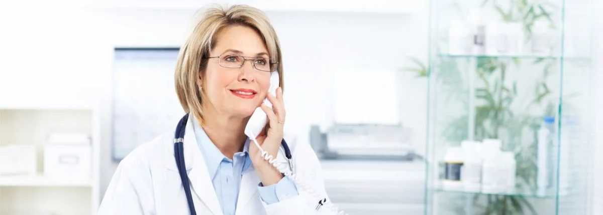 Ärztin mit Stethoskop telefoniert im hellen Behandlungsraum, im Hintergrund stehen Medizinprodukte im Regal.