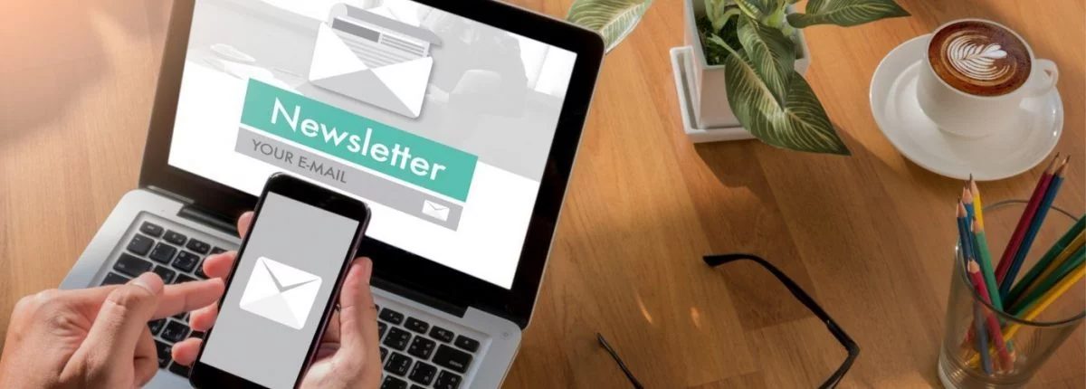 Eine Hand hält ein Smartphone mit E-Mail-Symbol vor einem Laptop mit Newsletter-Anmeldung auf einem Schreibtisch.