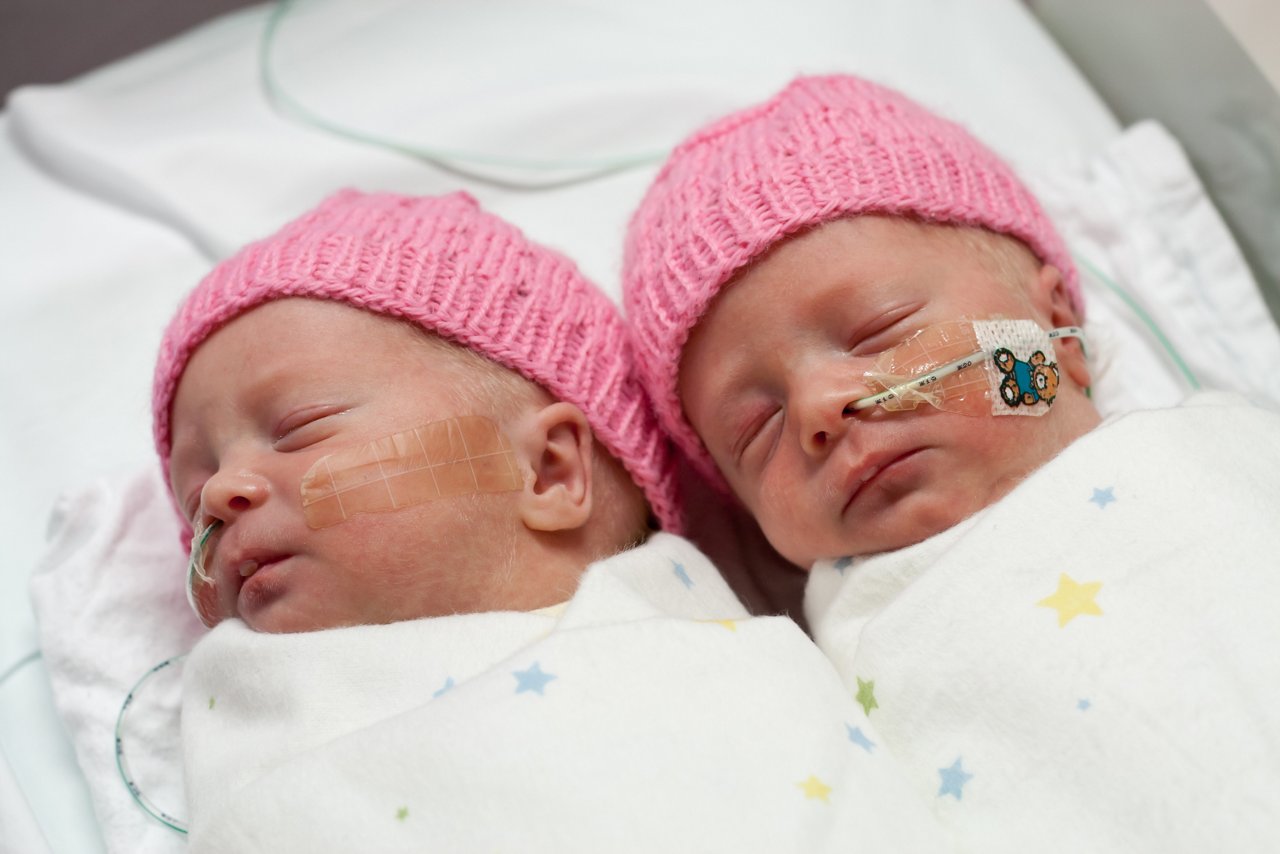 Newborn, premature twin girls asleep side by side in a special care nursery crib.