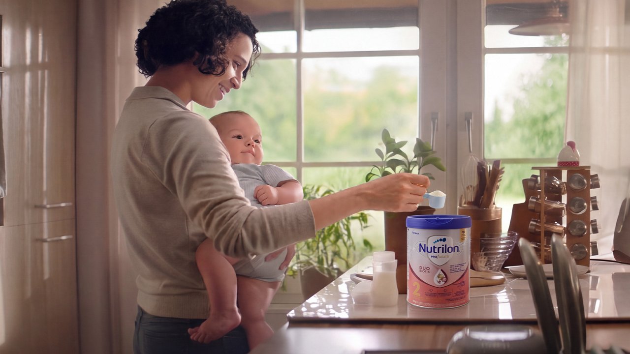 Persoon bereidt Nutrilon PROfutura babyvoeding in de keuken met een baby op de arm