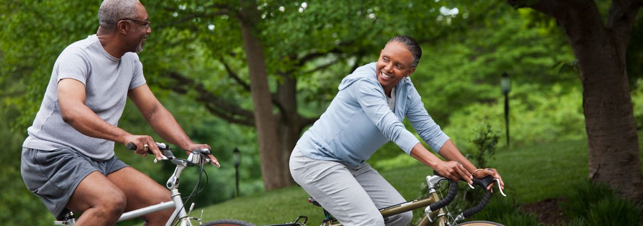 Dois indivíduos andam de bicicleta em um ambiente verde e cheio de árvores. Seus rostos estão obscurecidos para privacidade. A cena promove um estilo de vida ativo e saudável.