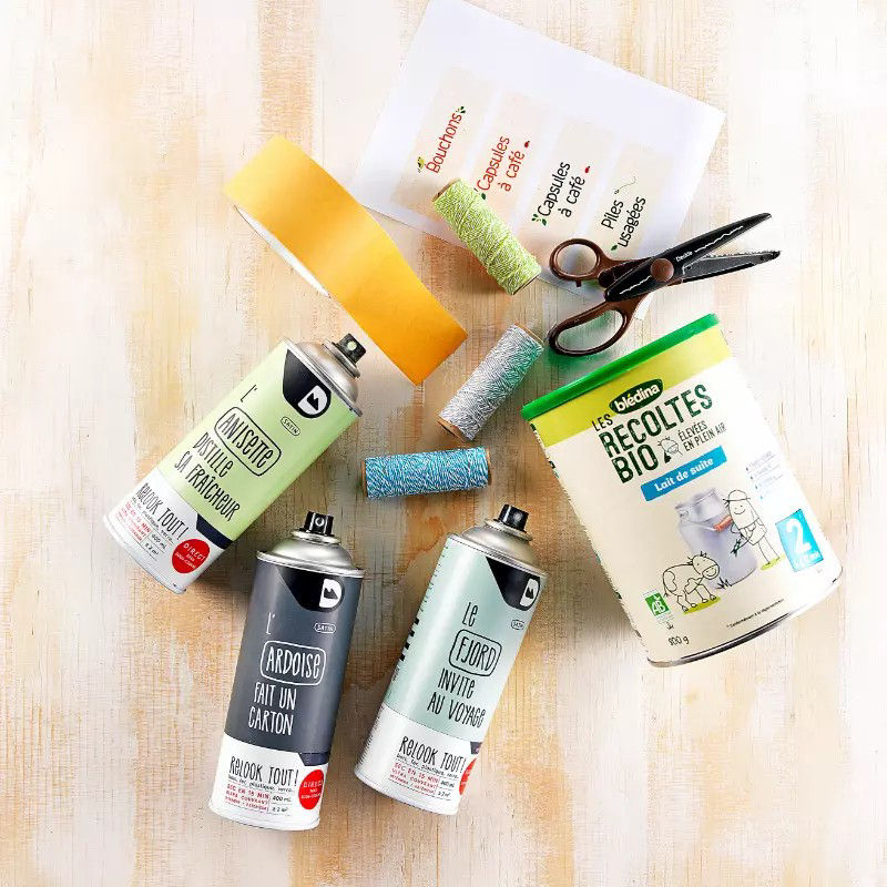 Une collection de fournitures artisanales comprenant des bombes de peinture, des rouleaux de ficelle coloree, des ciseaux et du ruban adhesif disposes sur une surface en bois clair. Le texte visible sur les bombes de peinture comprend des expressions telles que � Ardoise � et � Le Fluoro �, tandis que le conteneur vert de l' e affiche � Recoltes Bio � et � 500 g �. Le decor suggere une activite creative de bricolage ou d'artisanat.