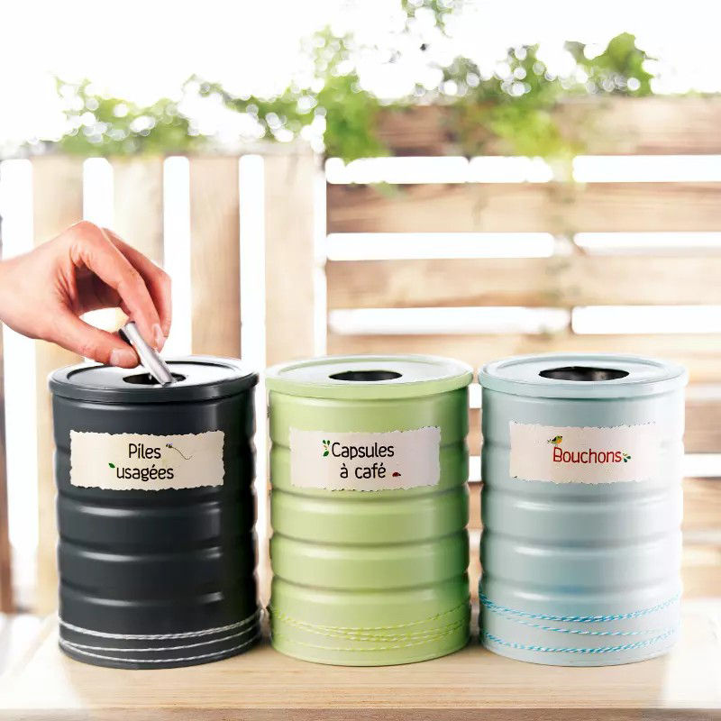 Trois poubelles de recyclage cylindriques sont disposees sur une surface en bois, etiquetees pour des types de dechets specifiques : � Piles usagees �, � Capsules a cafe � et � Bouchons �. On voit une main deposer un objet dans la poubelle noire. Le decor represente un environnement exterieur lumineux avec des panneaux en bois et de la verdure en arriere-plan.
