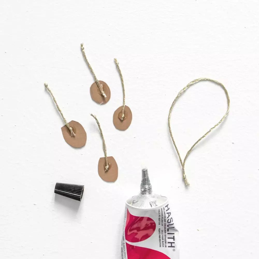 Un tube de colle Hasulith ouvert est presente sur une surface blanche a c�te de petites etiquettes marron auxquelles est attachee de la ficelle. Le tube de colle presente un motif rose et blanc avec du texte visible. La scene suggere une activite de bricolage ou d'artisanat impliquant des materiaux adhesifs et decoratifs.