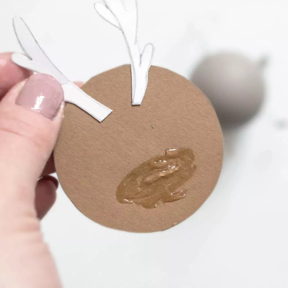 Gros plan sur une decoration de No�l artisanale representant un renne, avec une base circulaire marron et des bois en papier blanc. La decoration est tenue par une main aux ongles vernis de rose p�le, sur un fond neutre flou. Les traces de colle visibles ajoutent un charme rustique et artisanal au design.