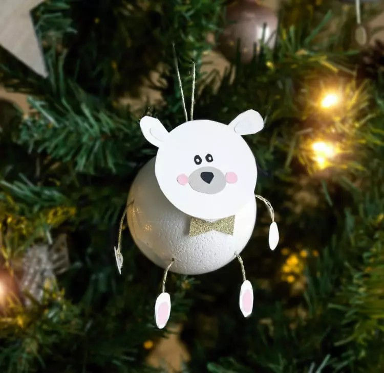 Une decoration artisanale en forme d'ours polaire est suspendue a un sapin de No�l decore. La decoration presente un corps spherique blanc, un visage d'ours souriant et des bras et des jambes pendants. Le decor festif comprend des lumieres scintillantes et des branches de pin vertes, creant une atmosphere chaleureuse de fetes de fin d'annee.